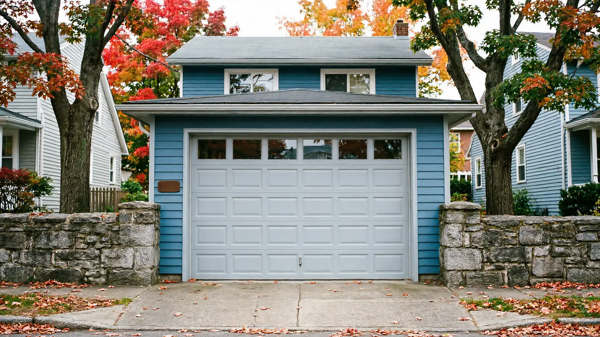A light gray garage door with windows is on a blue house in autumn.